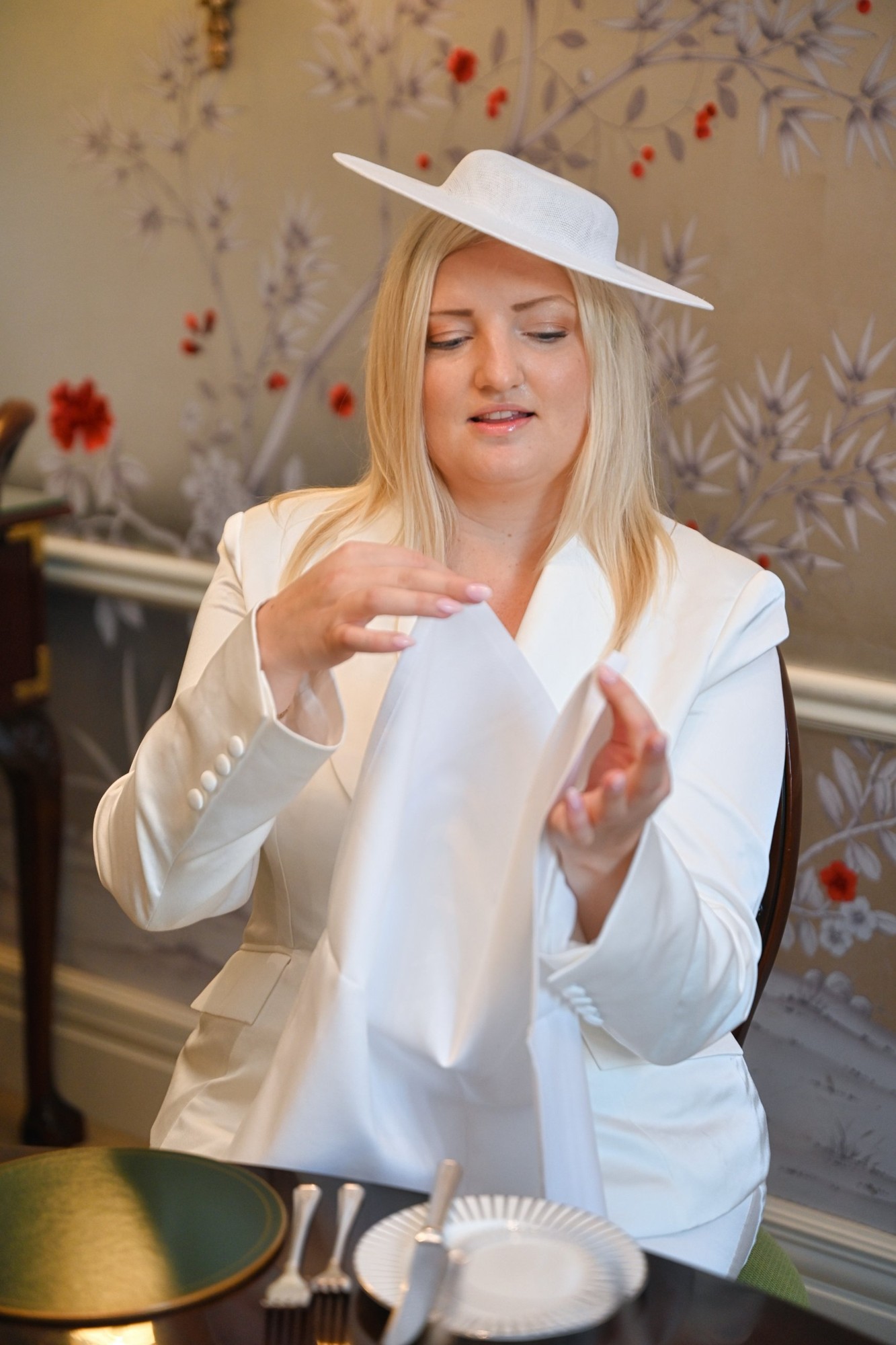 A woman in a white suit and hat unfolding a napkin at the table