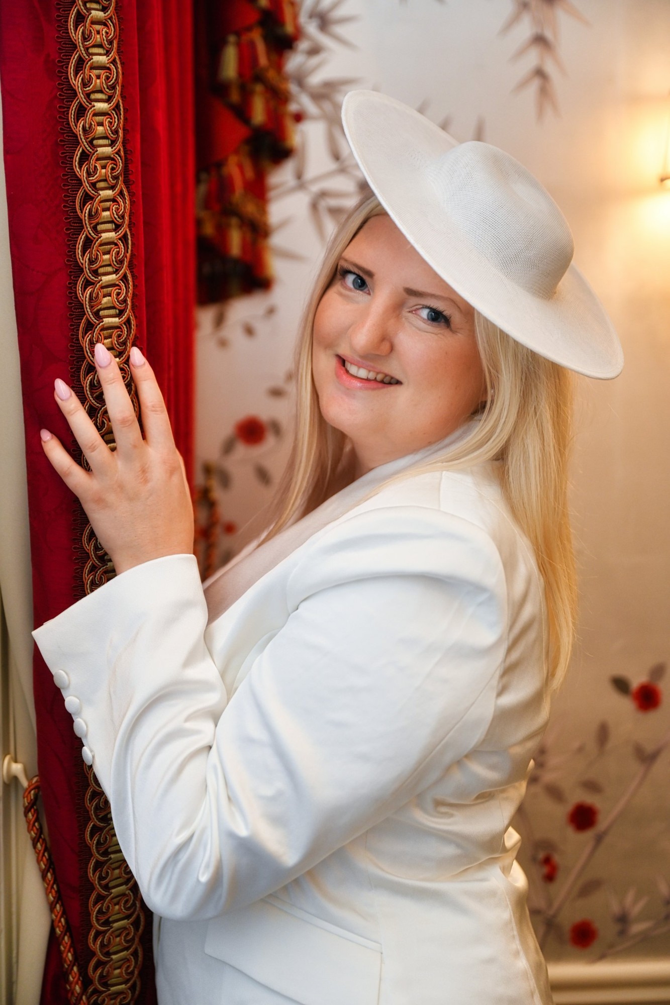 A smiling woman in a white suit and hat beside a red velvet curtain