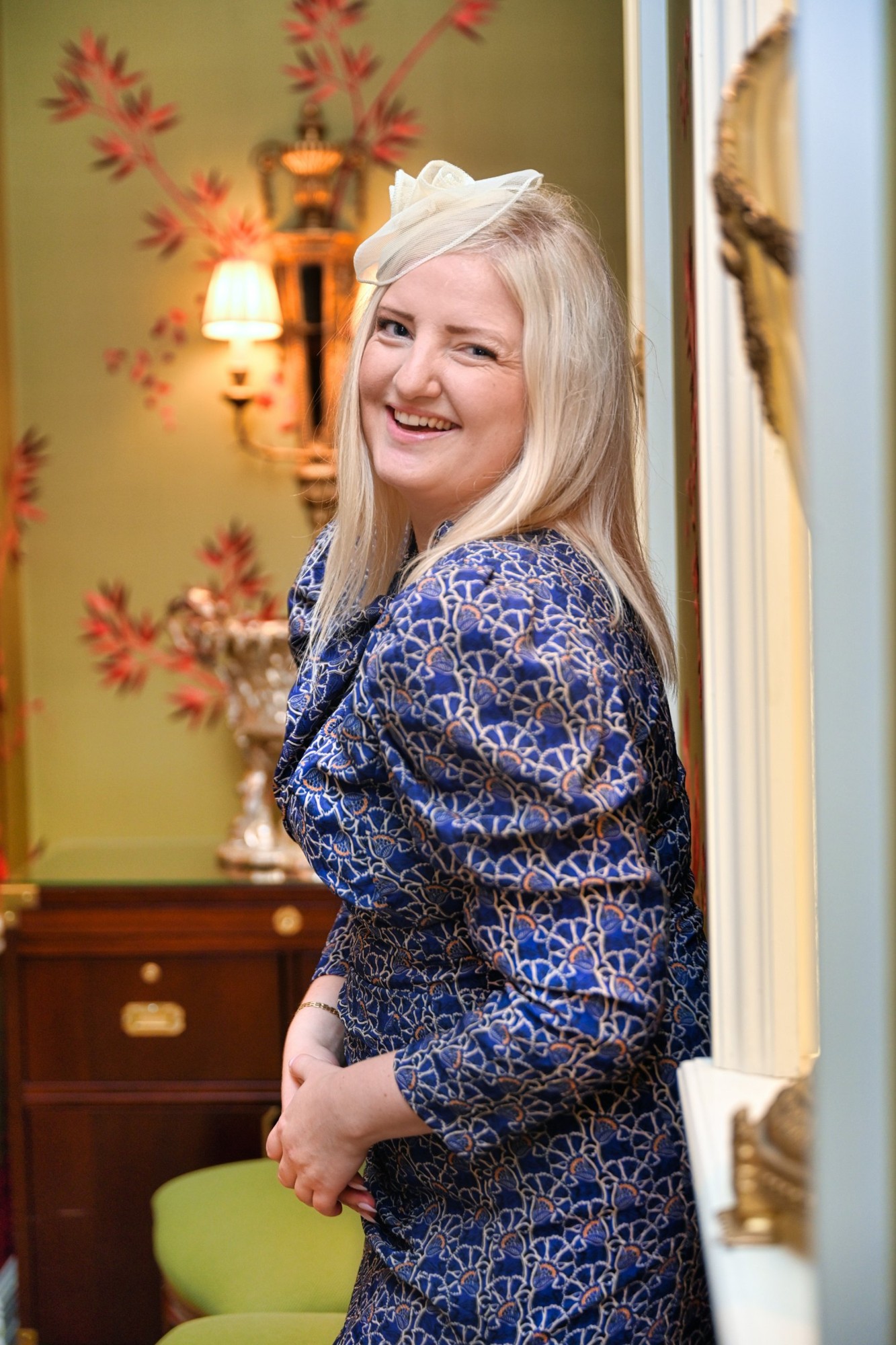 A laughing woman in a blue patterned dress and fascinator in a drawing room