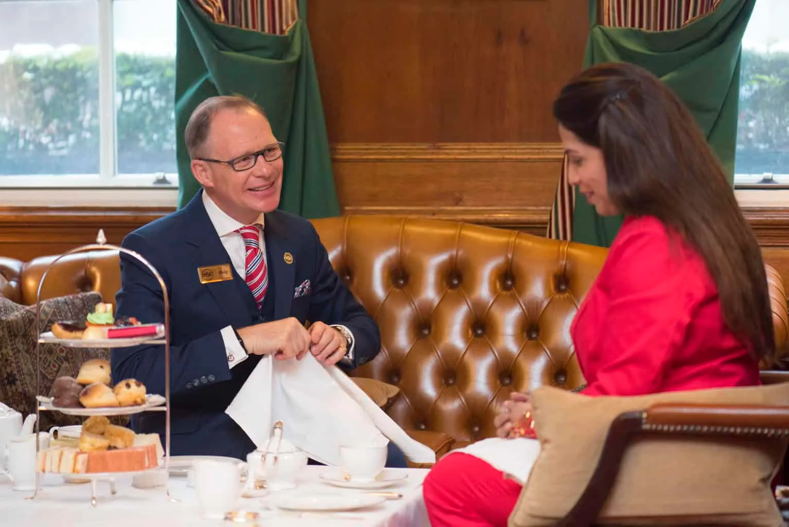 About 6 | The British School of Excellence | About Afternoon tea etiquette lesson. Philip sits at a table with lady in red dress.